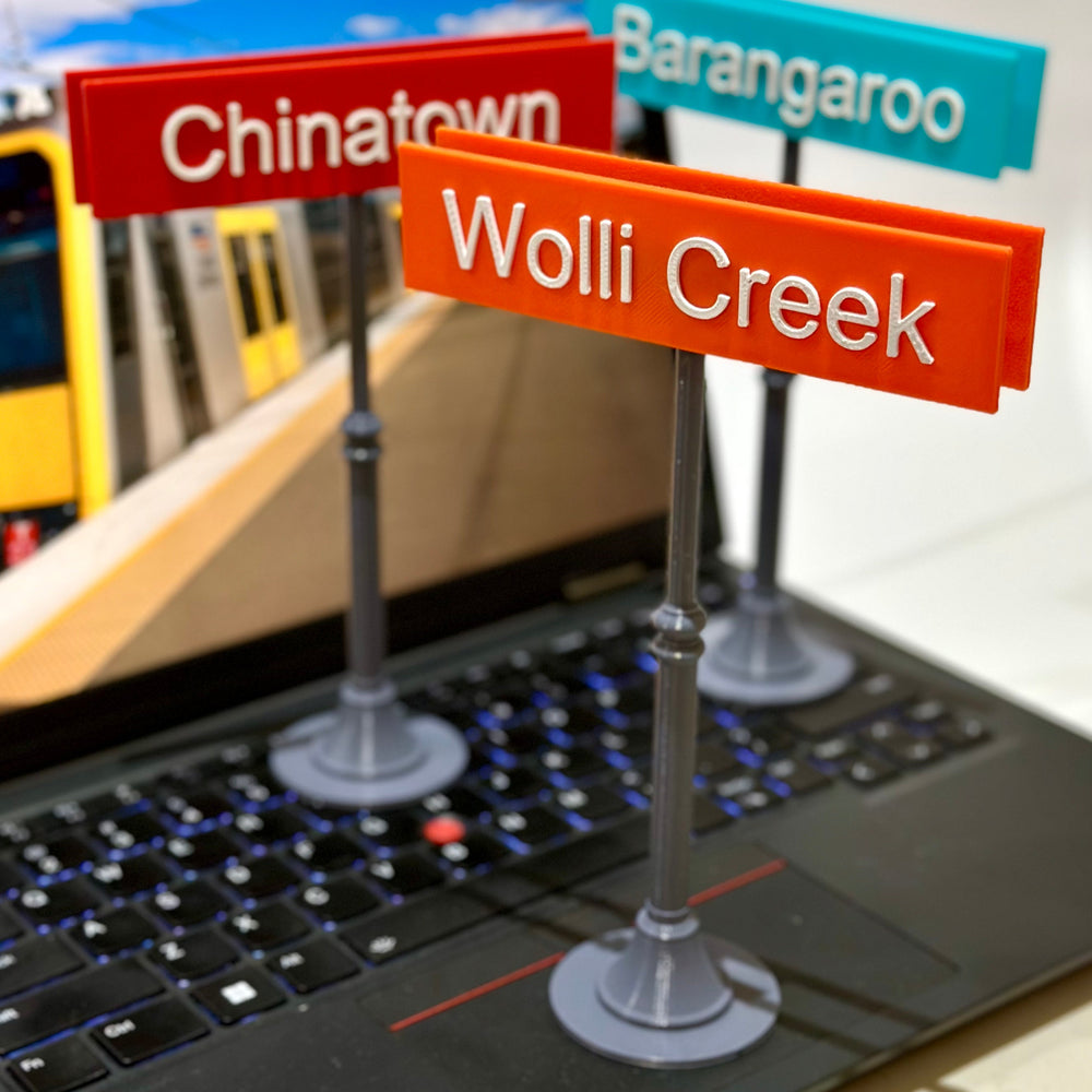 3D printed Sydney station signs colour-coded by mode — Trains orange, Metro blue, Light Rail red — showing Chinatown, Barangaroo and Wolli Creek